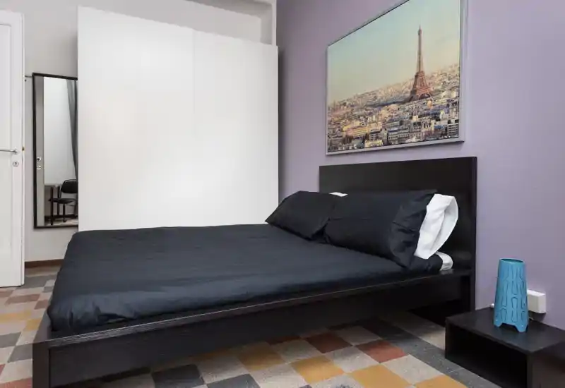 Bright, modern bedroom with a large dark-wood bed dressed in black linens, bedside table and decorative wall art showing the Eiffel Tower. The room is clean, well-lit and suitable for rental photos.