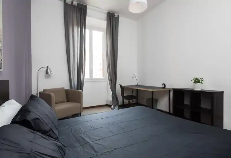 Bright, modern bedroom with a large bed dressed in dark linens, a comfortable armchair, desk workspace and shelving near a window that provides natural light.