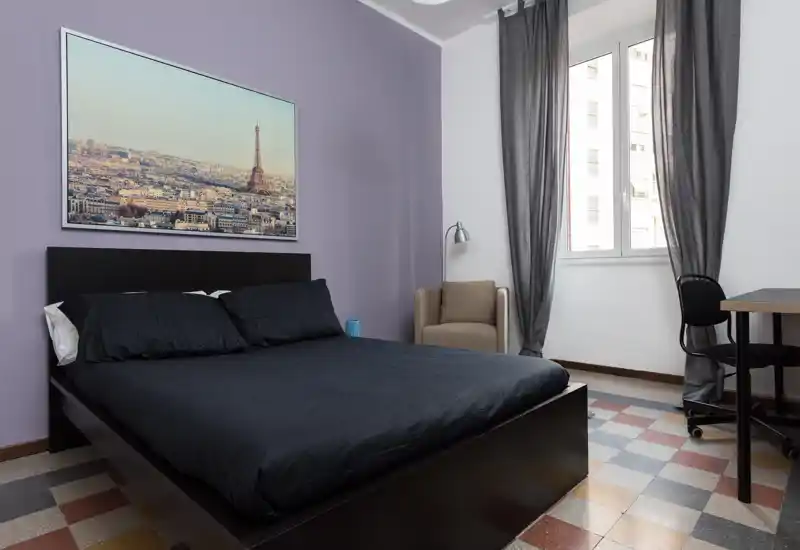 Bright, modern bedroom with a queen bed dressed in dark linens, an accent wall with framed artwork, a comfortable armchair, desk and natural light from a large window.