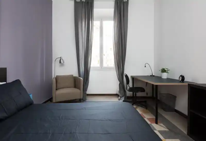Bright, minimally styled bedroom with a large bed in the foreground, a comfortable armchair by the window and a practical desk workspace — ideal for students or professionals.