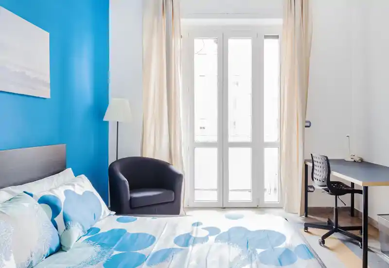 Camera da letto luminosa e moderna con ampia finestra, biancheria da letto blu a fantasia e una poltroncina comoda — perfetta per un soggiorno accogliente.