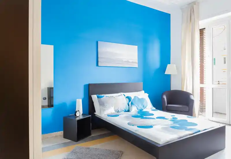 Bright, modern bedroom with a blue feature wall, double bed, bedside table and natural light from a balcony door — clean and inviting for guests.