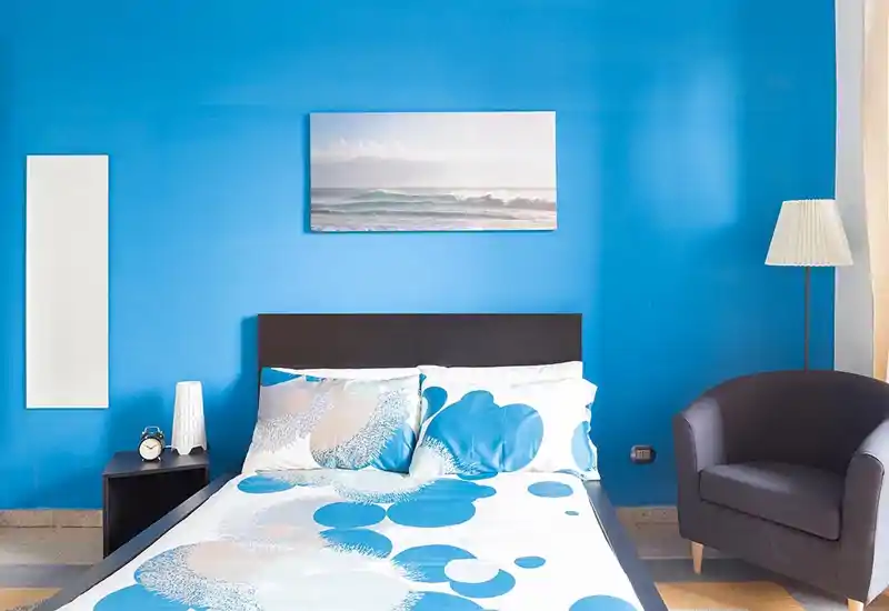 Bright, modern bedroom with a queen bed against a bold blue wall, patterned bedding, bedside table with lamp and an armchair — well-lit and stylish.