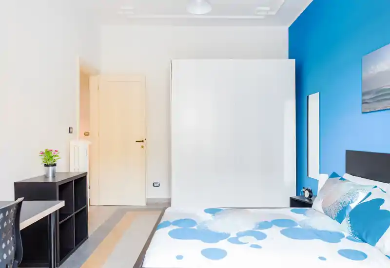 Bright, modern bedroom with a blue accent wall, double bed with coordinated bedding, large white wardrobe and simple desk — ideal for a clean, stylish rental.