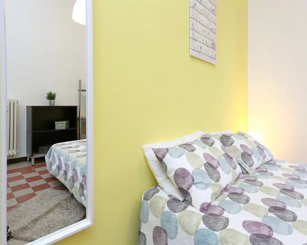 Camera da letto luminosa e ordinata con biancheria a fantasia, cuscini e una lampada da comodino calda; parete gialla d'accento e uno specchio che riflette parte della stanza.