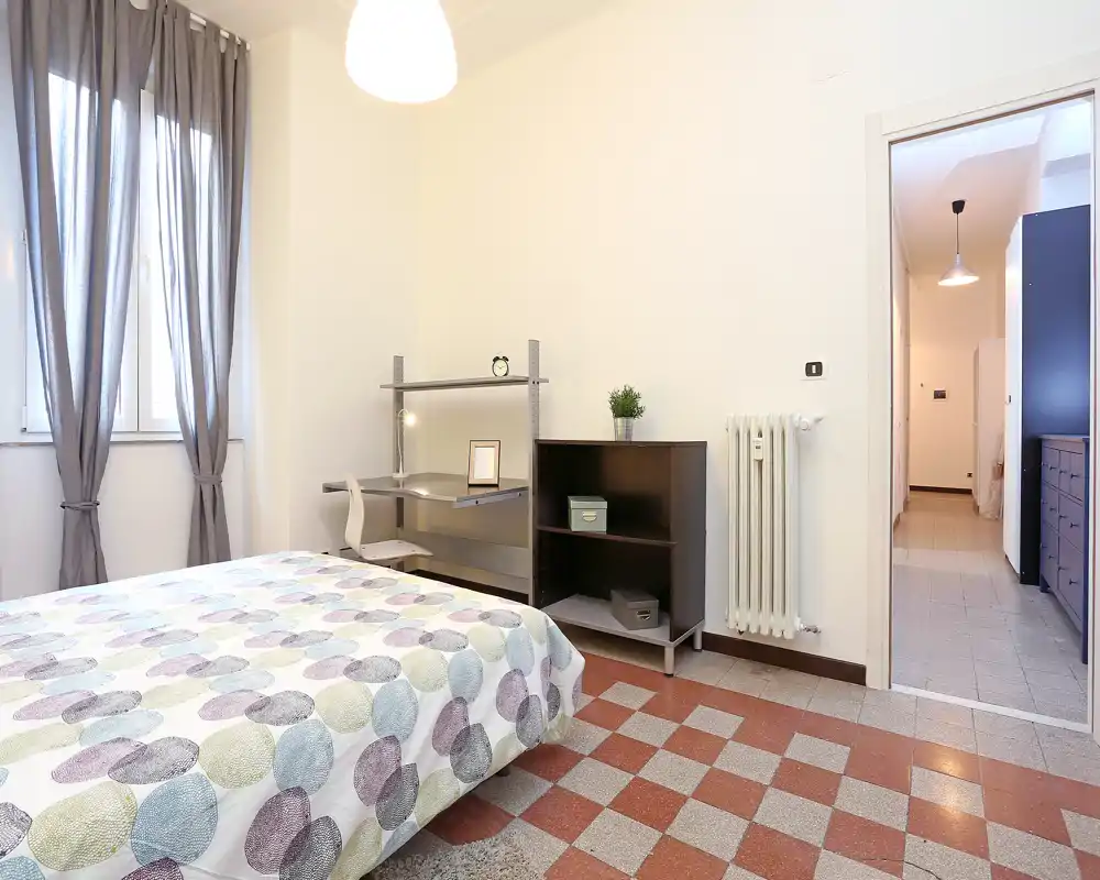 Camera da letto luminosa e ben arredata con letto matrimoniale a fantasia, angolo scrivania e mobile contenitore. Ampia finestra con tende per luce naturale e pavimento piastrellato che caratterizza l'ambiente.