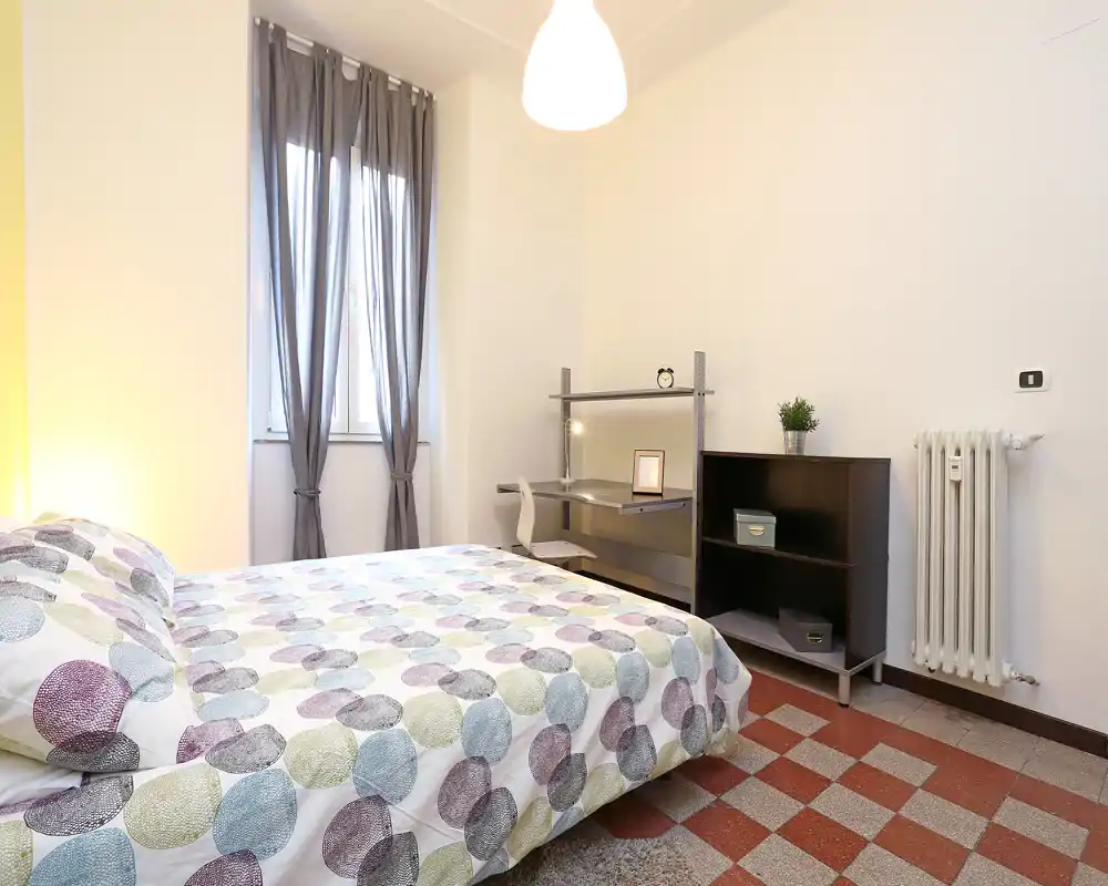 Camera da letto luminosa e ordinata con letto matrimoniale, biancheria a fantasia, scrivania e scaffale; luce naturale dalla finestra con tenda e lampada centrale creano un’atmosfera accogliente.