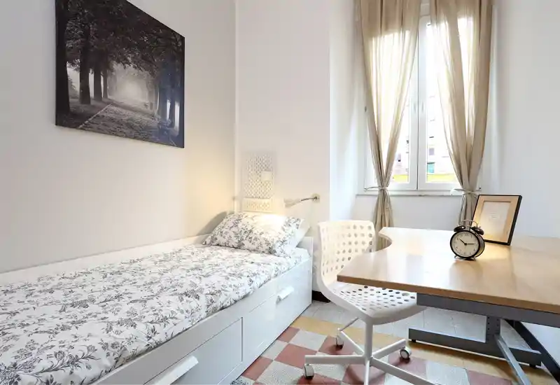 Small, well-lit single bedroom with a neatly made bed, study desk and natural light from a large window. Clean, modern styling with soft textiles and a framed wall picture.