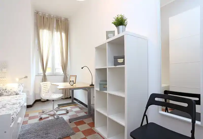 Bright, tidy bedroom with a single bed, a compact workspace by the window, shelving unit and minimalist décor — ideal for a student or single occupant.