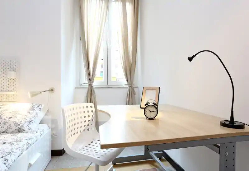 Bright, minimalist bedroom corner with a neatly made bed, a wooden desk, modern chair and natural window light — ideal for a calm workspace and rest.