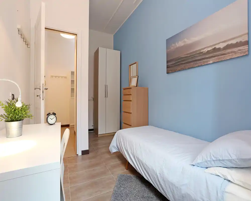 Una camera singola ordinata con letto ben rifatto, parete azzurra, armadio e comò — luminosa, tranquilla e ben allestita per affitti.