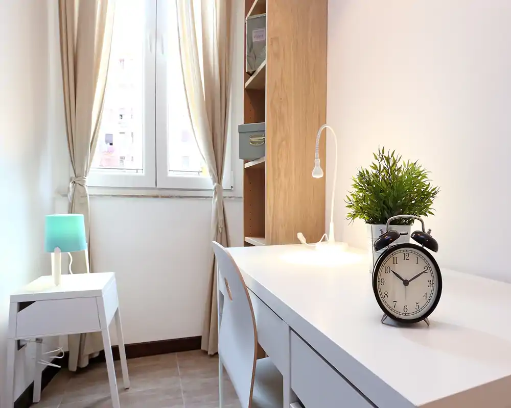 Spazioso angolo camera luminoso e minimale con scrivania bianca, comodino e luce naturale dalla finestra. La scrivania ordinata con orologio vintage e pianta rende la stanza ideale per una camera singola compatta.