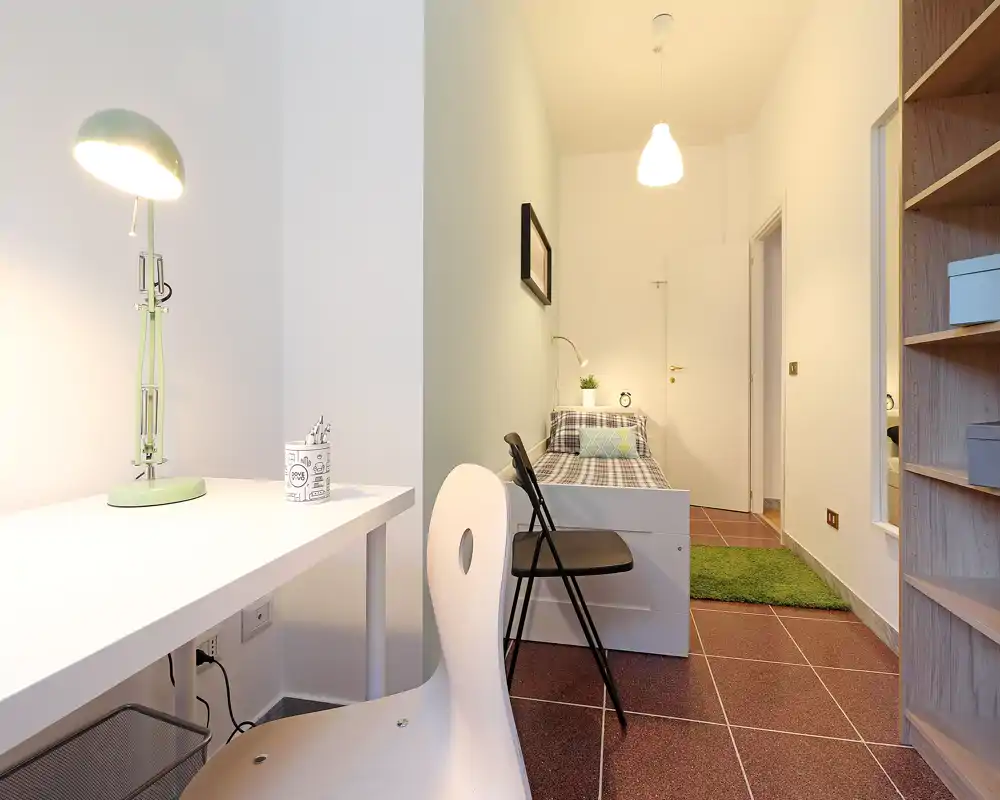 Camera luminosa e ben arredata con una scrivania in primo piano e un letto singolo ordinato con lampada da comodino, ideale per uno studente o un occupante singolo.