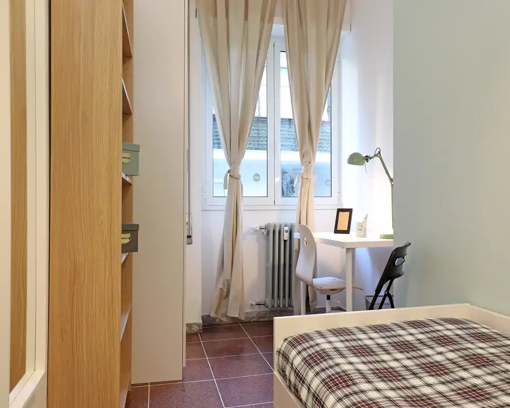 Camera da letto luminosa e ordinata con letto singolo in primo piano, armadio a sinistra e scrivania vicino alla finestra — ideale per uno studente o una persona sola.