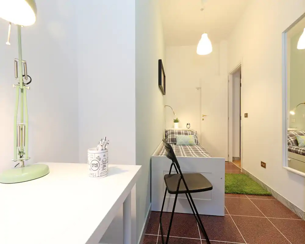 Piccola camera singola ben illuminata con letto ordinato, lampada da scrivania e arredo moderno minimalista — ideale per studenti o soggiorni brevi.