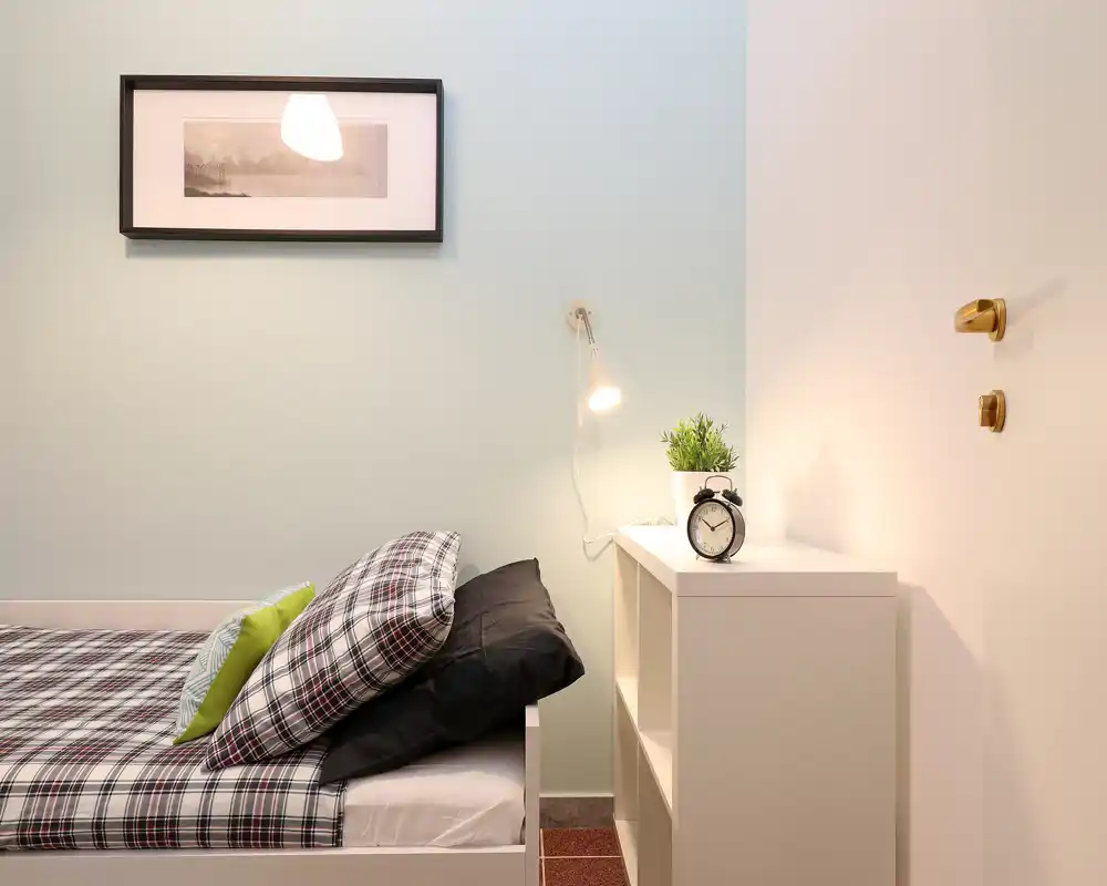 Angolo camera da letto compatto e curato con letto singolo in lenzuola a quadri, comodino a ripiani, lampada da lettura e sveglia — luminoso, ordinato e accogliente.