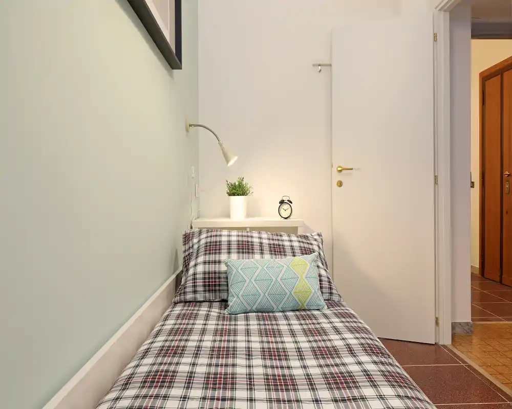 Camera singola luminosa e ben curata con copriletto scozzese, cuscino decorativo, mensola notte con lampada, pianta e sveglia — perfetta per un singolo o ospiti.