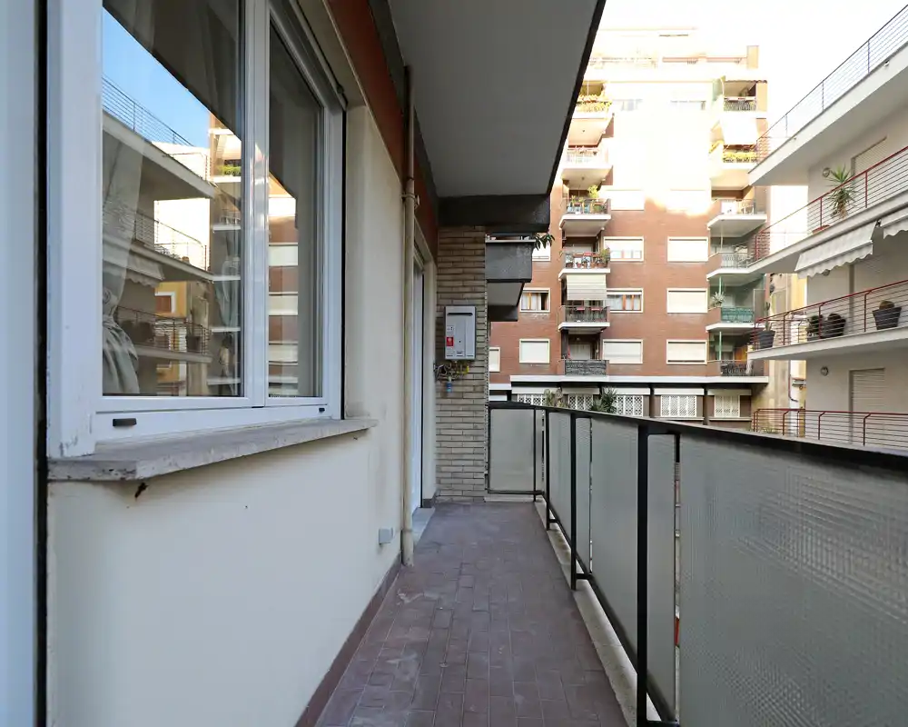 Balcón largo y estrecho con barandilla de vidrio esmerilado, suelo de baldosas y vistas a los edificios vecinos; apto como zona exterior para sentarse.