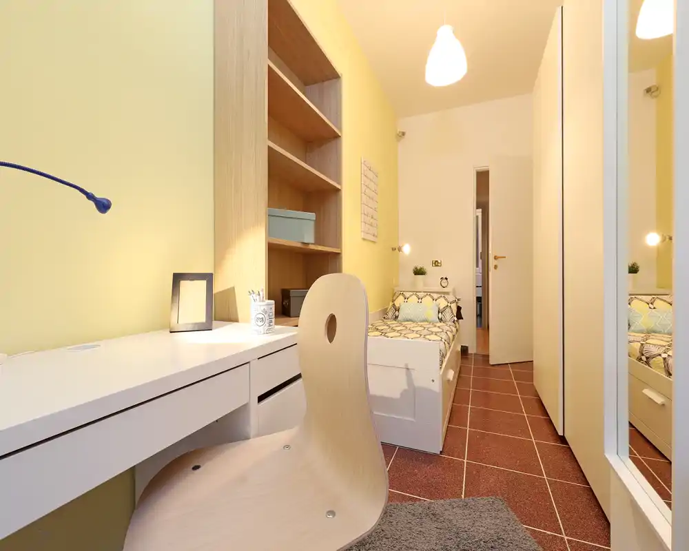 Dormitorio individual compacto con escritorio integrado, estanterías y una cama sencilla ordenada; paredes amarillas cálidas e iluminación suave crean un espacio acogedor y bien organizado, ideal para un estudiante o una sola persona.