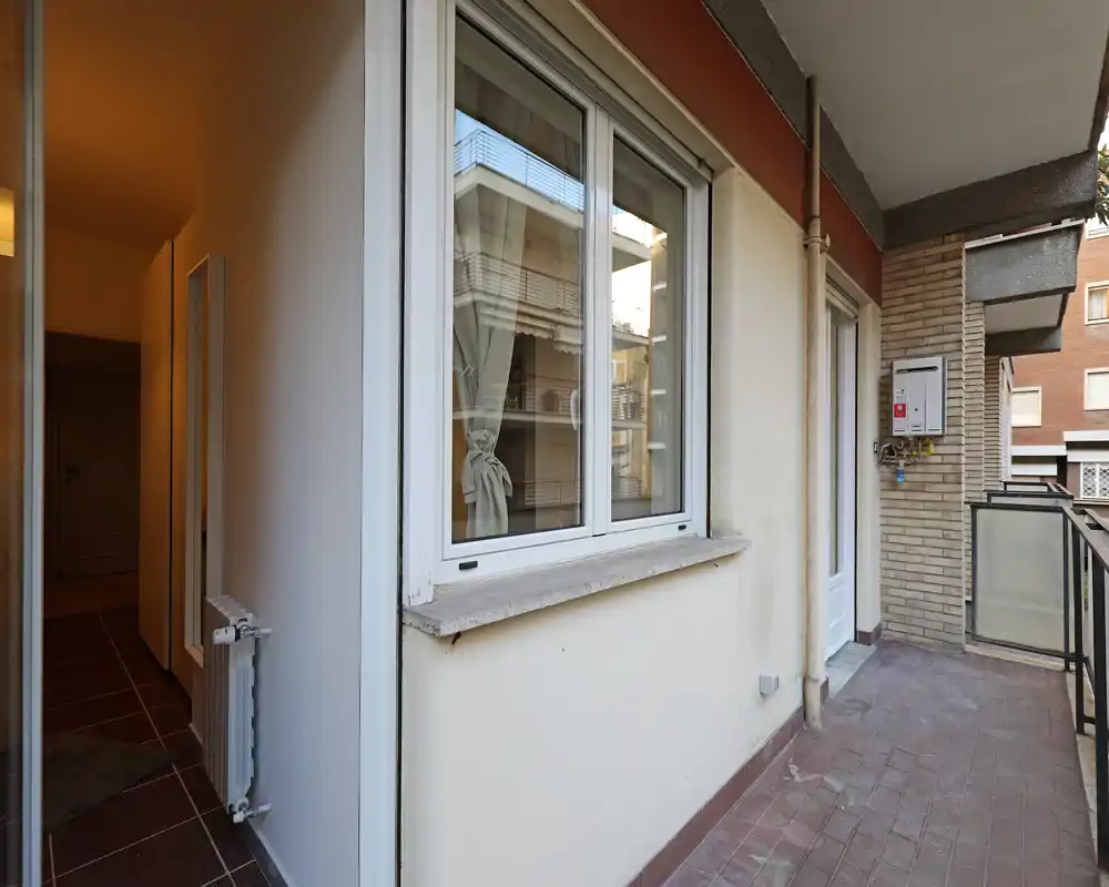 Balcón exterior estrecho con suelo de baldosas, barandilla y puerta de acceso al interior. El espacio es adecuado para pequeños muebles o plantas y muestra la fachada del edificio y ventanas vecinas.