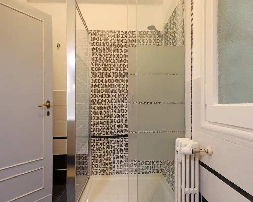Bagno moderno e pulito con box doccia in vetro, piastrelle a mosaico e termosifone a parete accanto a una finestra con vetro satinato.