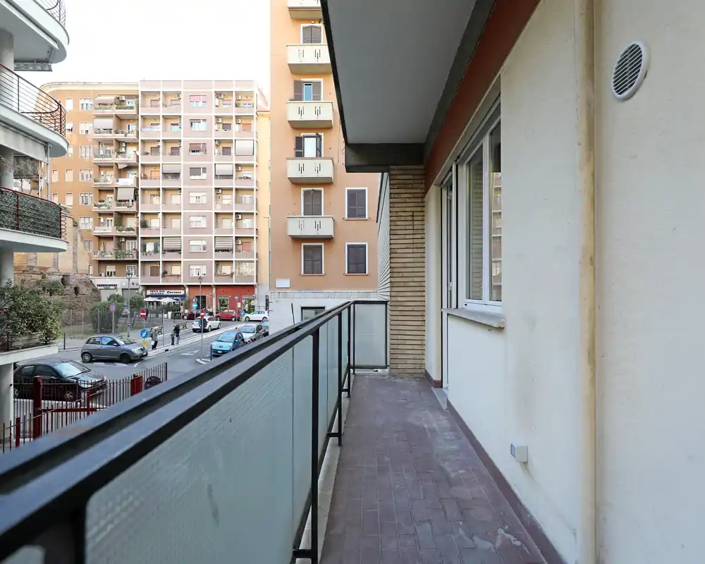 Balcone rivolto sulla strada con parapetto in vetro satinato, pavimento piastrellato e accesso dall'appartamento; ottima luce naturale e vista urbana.