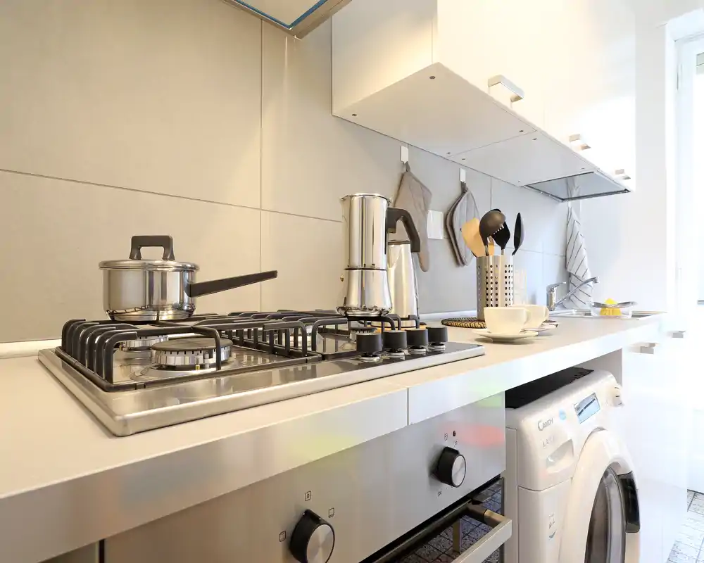 Cucina moderna e luminosa con piano cottura a gas in acciaio inox, forno compatto e lavatrice integrata, con accessori sul piano ordinati.