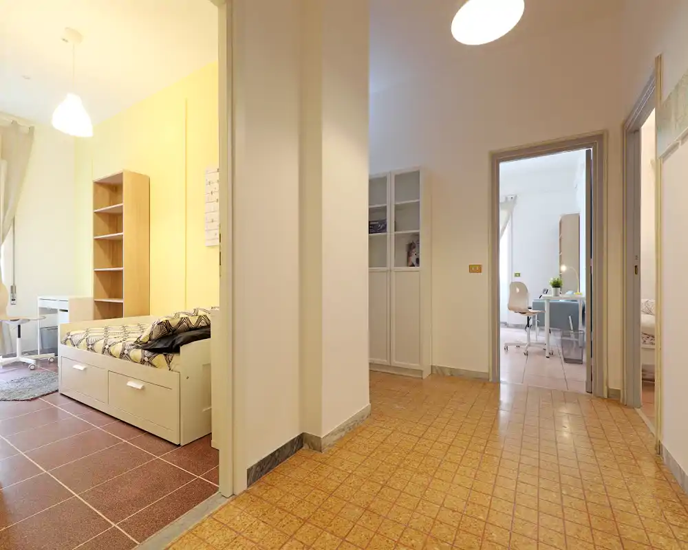 Angolo camera luminoso e ordinato con letto singolo, scaffale e scrivania visibili attraverso la porta aperta. Parete gialla e tessili creano un’atmosfera accogliente, perfetta per annunci di affitto.