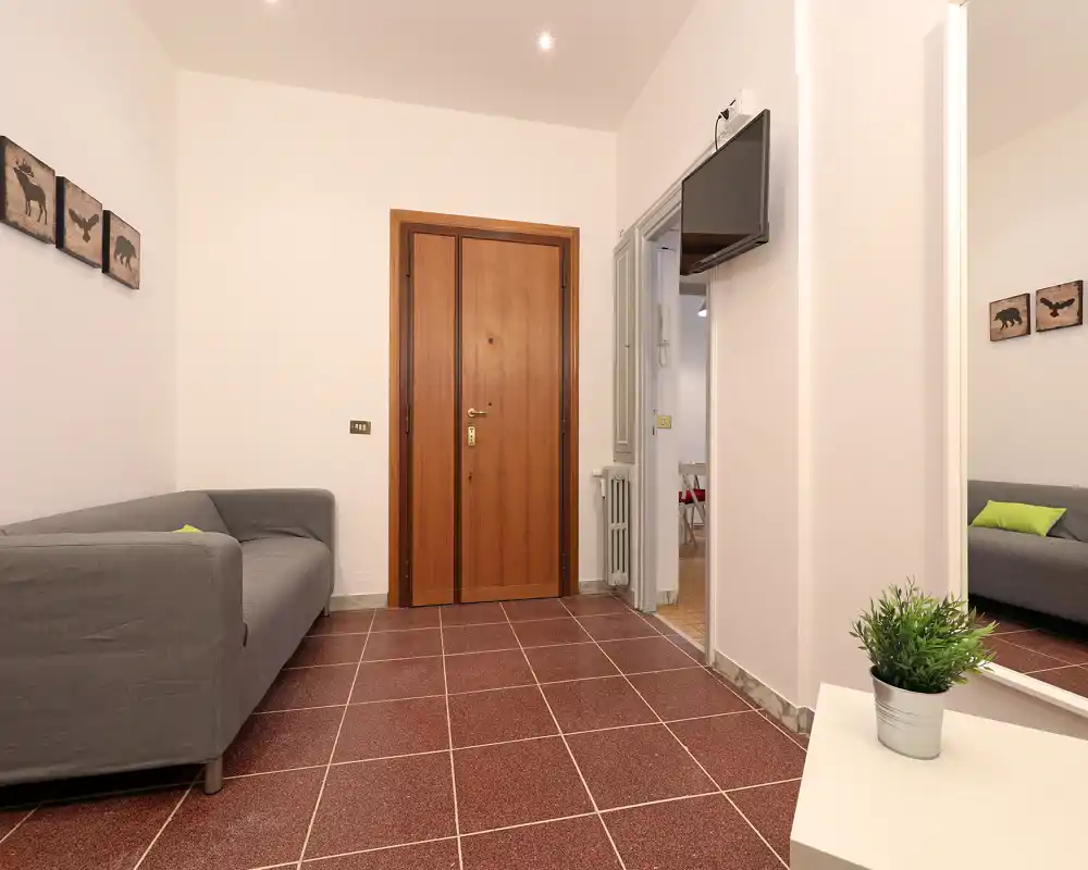 Piccolo ingresso ordinato con porta d'ingresso in legno, divano grigio, TV a parete e uno specchio grande che riflette l'area salotto. Pareti neutre e pavimento caldo donano una prima impressione accogliente.