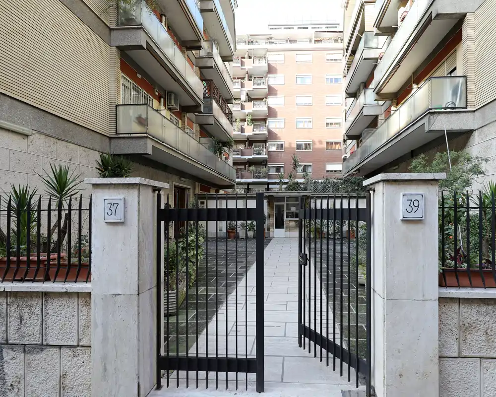 Ingresso recintato dell'edificio che conduce a un cortile centrale con balconi e vasi — ben illuminato e curato.