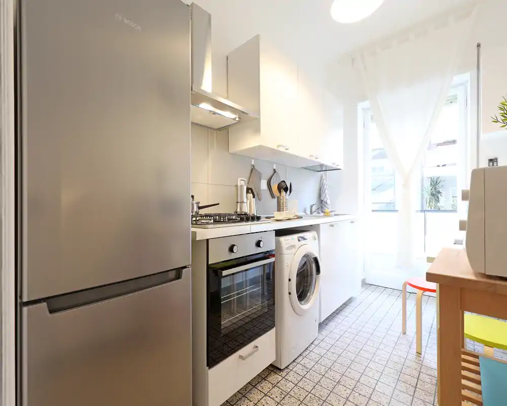 Cucina moderna e luminosa con frigorifero in acciaio, forno incassato, piano cottura a gas e lavatrice; mobili bianchi e finestra soleggiata per un ambiente pulito e arioso.