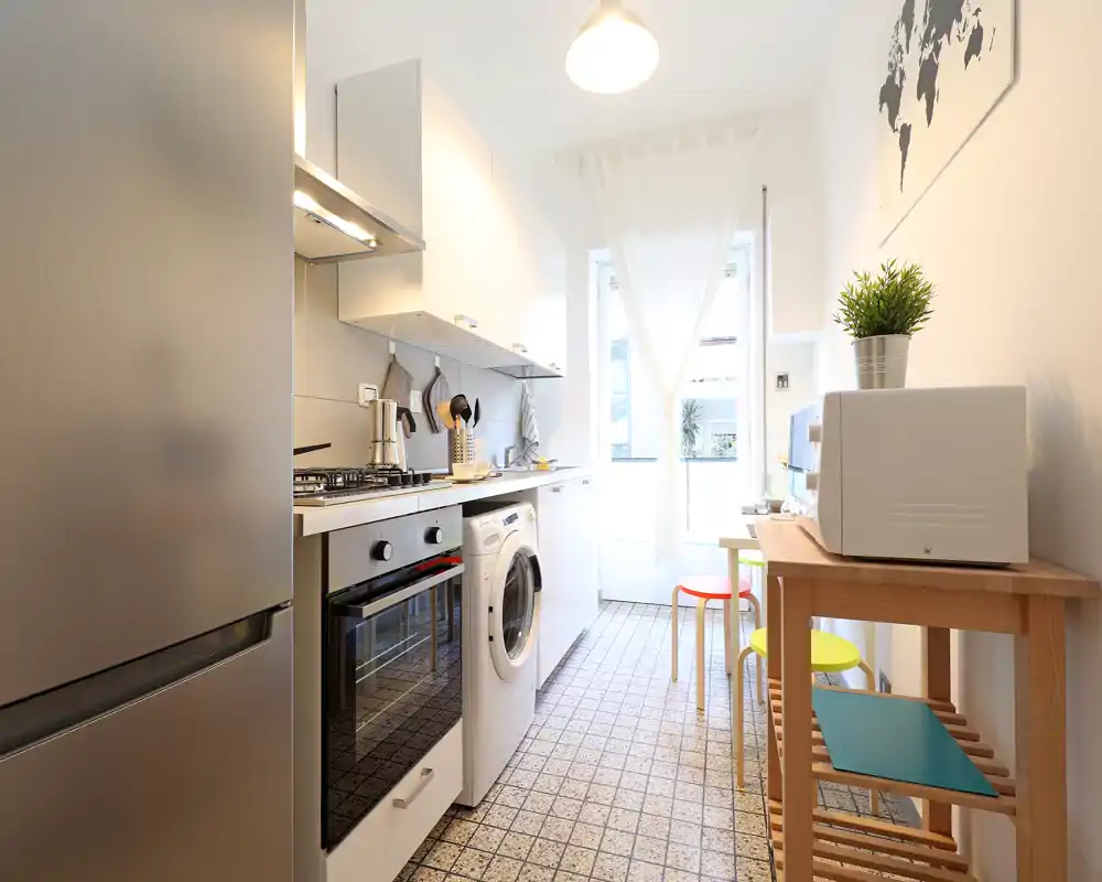 Cucina moderna a corridoio, luminosa, con elettrodomestici in acciaio, piano cottura a gas, forno incassato e lavatrice; piccolo angolo pranzo con sgabelli colorati e luce naturale.