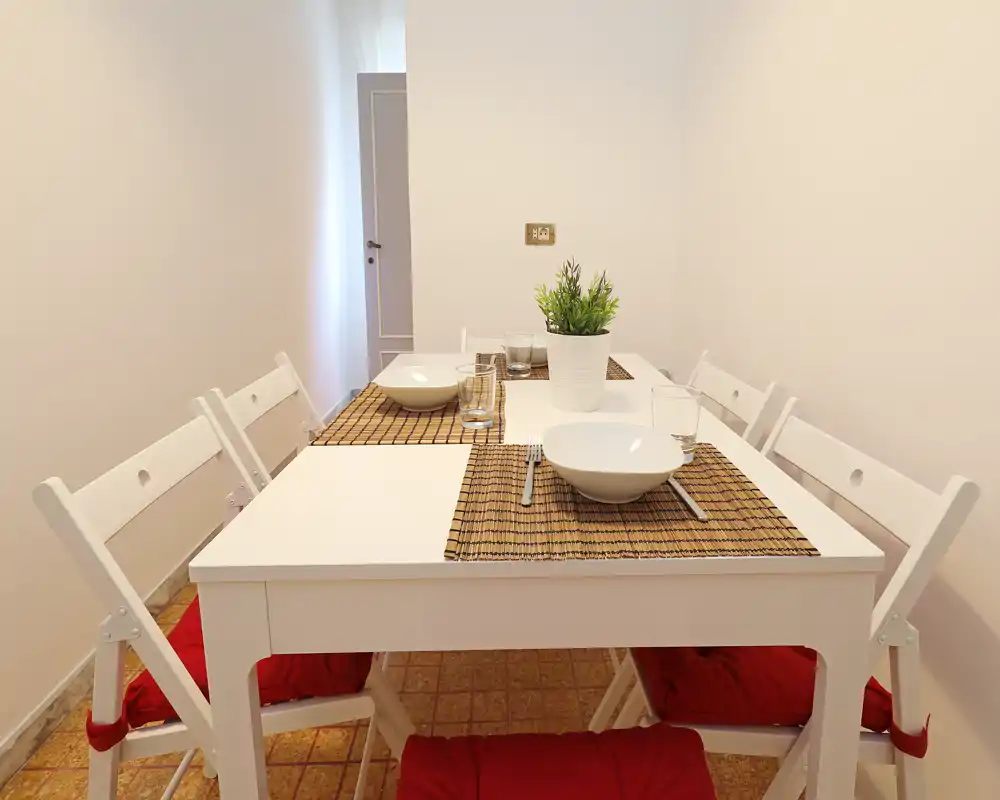 Zona pranzo luminosa e minimale con tavolo bianco per quattro, tovagliette intrecciate e cuscini rossi sulle sedie che creano un’atmosfera calda e ordinata.