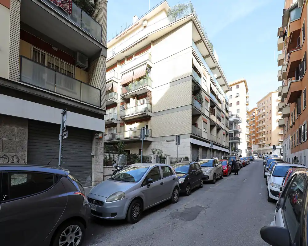 Vista di strada in pieno giorno con auto parcheggiate e palazzi residenziali su una strada urbana stretta, utile come contesto esterno/ubicazione.