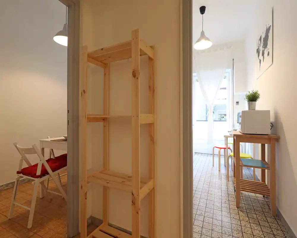 Zona giorno/pranzo compatta e luminosa con mobili in legno essenziali, un piccolo tavolo con sedie e scaffalatura; luce naturale che entra dalle tende leggere.