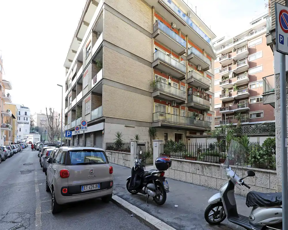 Vista esterna a livello strada di un edificio residenziale con balconi, auto e scooter parcheggiati—ideale per mostrare la facciata e il contesto di quartiere.
