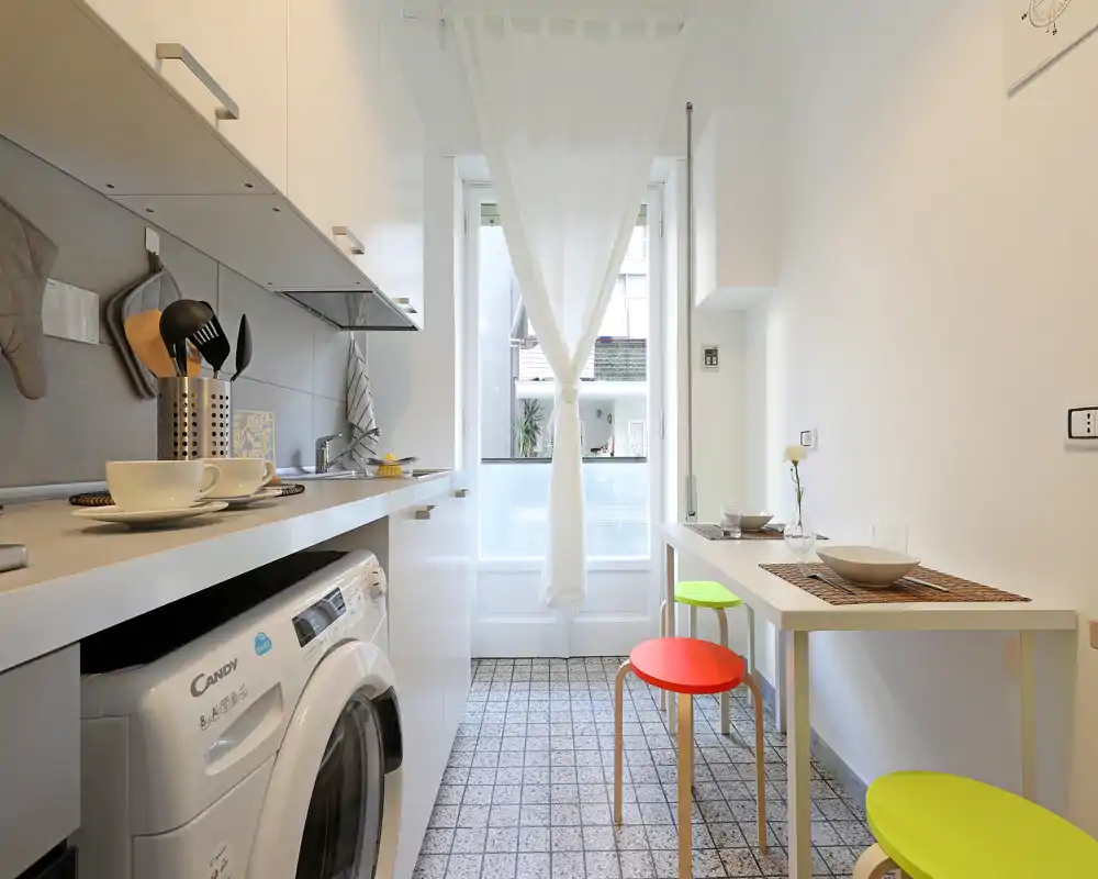 Cucina compatta e luminosa con piano di lavoro, lavatrice incorporata e piccolo tavolo da pranzo con sgabelli colorati — perfetta per un monolocale moderno.