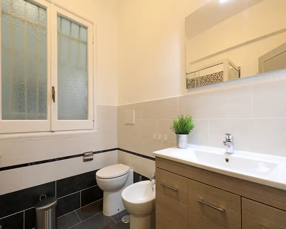 Bagno moderno e luminoso con lavabo, specchio, bidet e WC; piastrelle pulite e luce naturale da una finestra satinata.