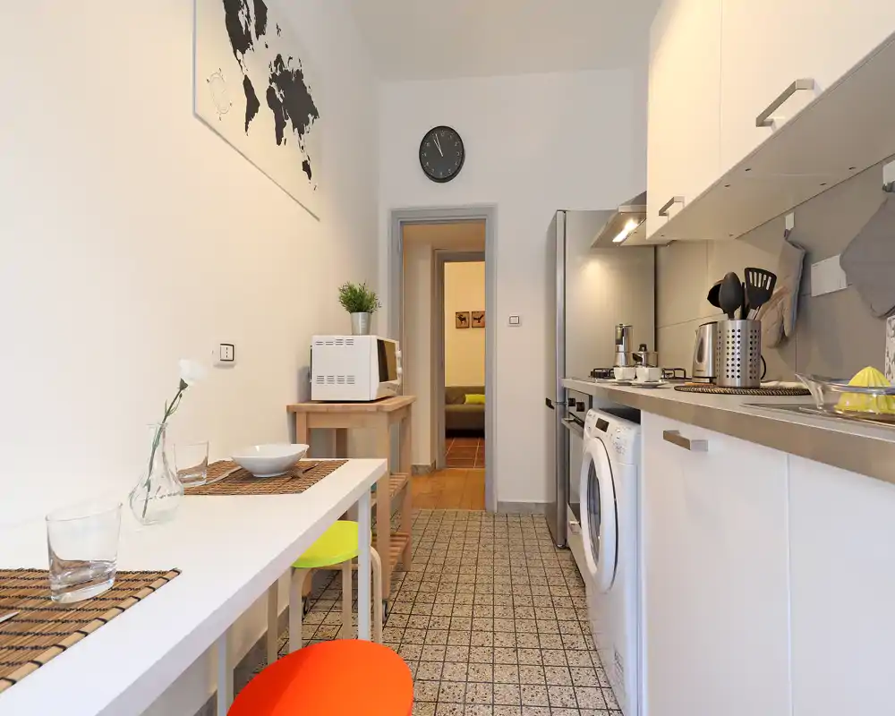 Cucina moderna e compatta con pensili bianchi, elettrodomestici in acciaio e un bancone pranzo stretto con sgabelli colorati. Ambiente luminoso, ordinato e dotato di essenziali come forno a microonde, frigorifero e piano cottura.