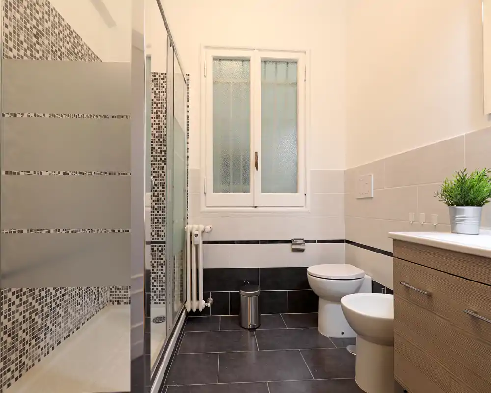 Bagno moderno e luminoso con doccia a parete in vetro, wc sospeso, bidet e mobile lavabo in legno. Accenti in mosaico e una finestra satinata garantiscono privacy lasciando entrare luce.
