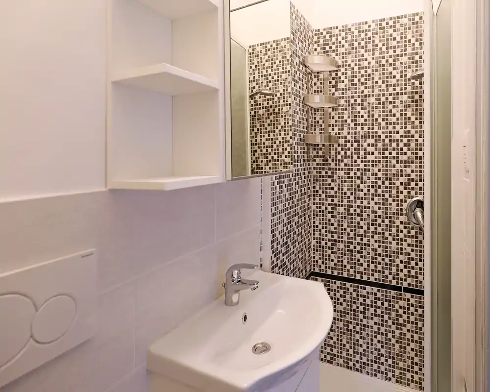 Bagno moderno e compatto con lavabo bianco e angolo doccia rivestito in mosaico; finiture pulite e specchio contenitore sopra il lavabo.