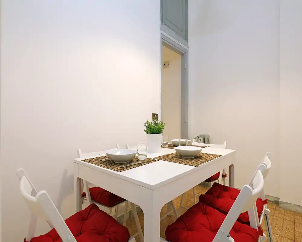 Zona pranzo moderna e luminosa con tavolo bianco per quattro, sedie con cuscini rossi e stoviglie essenziali — uno spazio compatto e accogliente per i pasti.