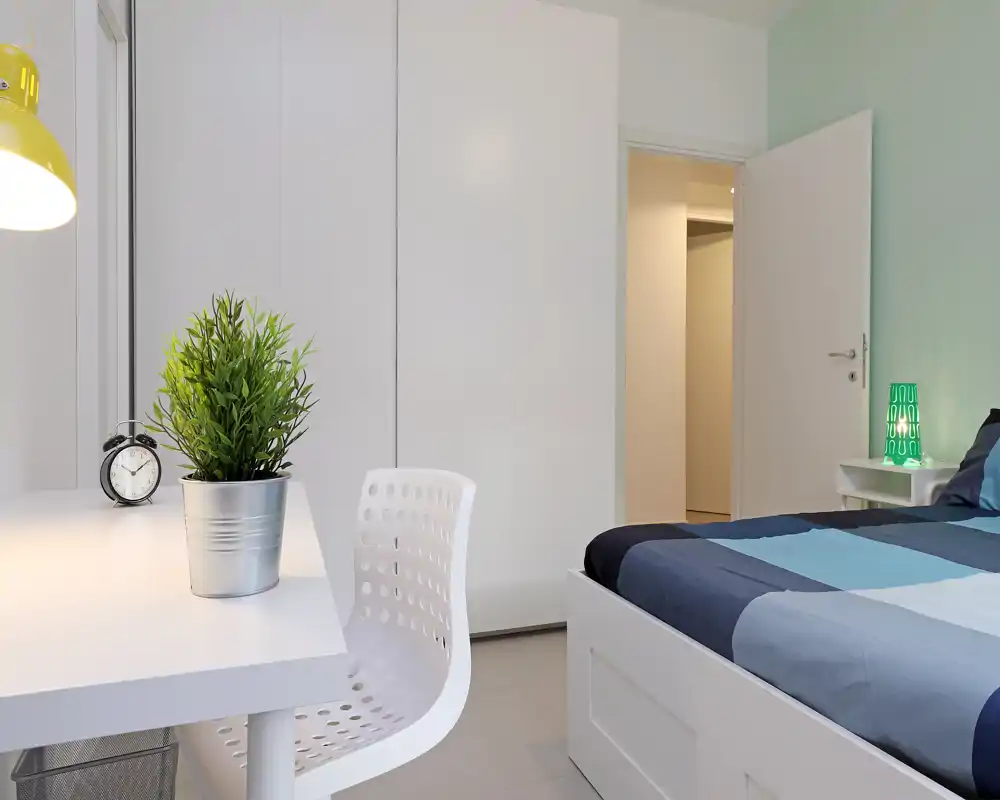 Bright, modern bedroom with a neatly made double bed, bedside table with lamp, built-in wardrobe and a simple desk area—ideal for a restful stay or student accommodation.