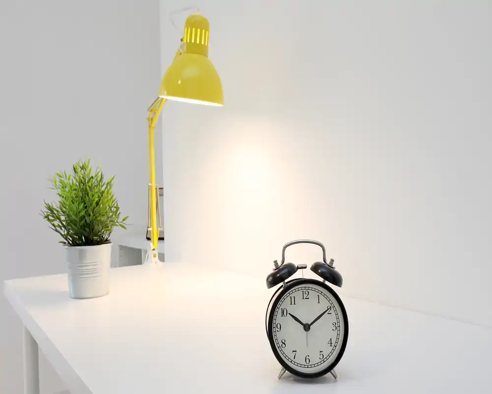 Minimal, well-lit white desk scene with a yellow desk lamp, small potted plant and a classic alarm clock on a clean surface.