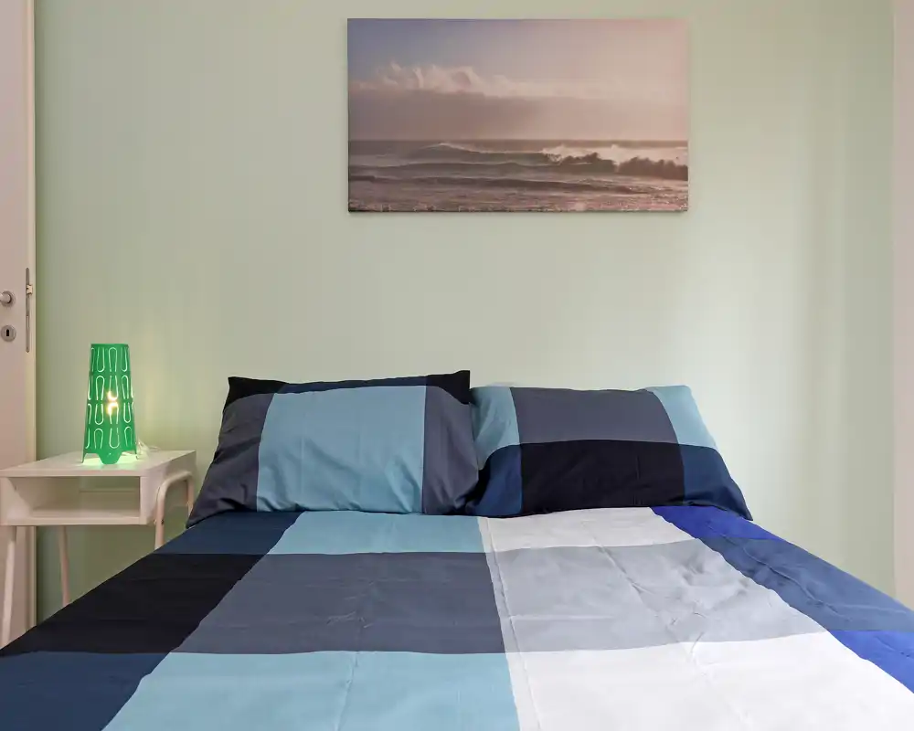 Bright, well-composed bedroom with a large double bed featuring blue check bedding, bedside table and a decorative lamp; calm wall art above the headboard enhances the relaxing atmosphere.