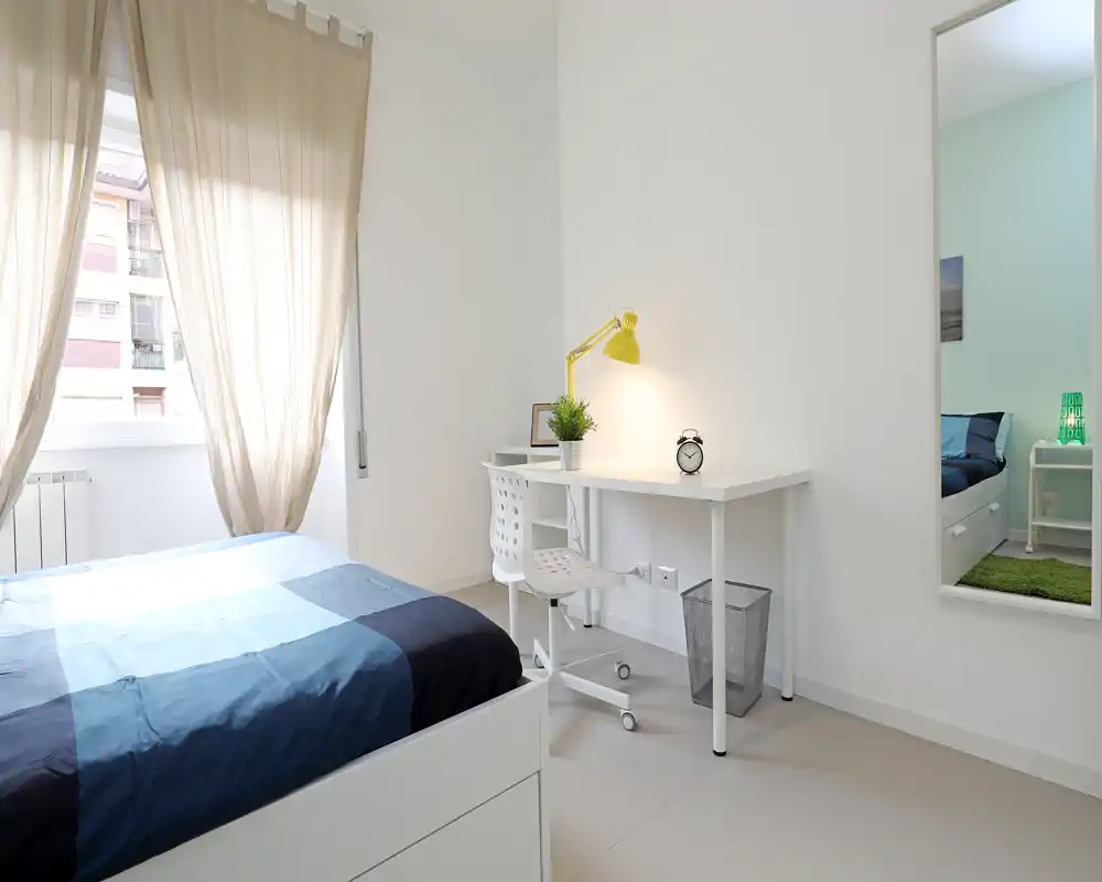 Bright, modern bedroom with a large window, simple white desk and a neatly made bed with blue linens. The room feels airy and well-lit, ideal for rental photos highlighting clean lines and a study corner.