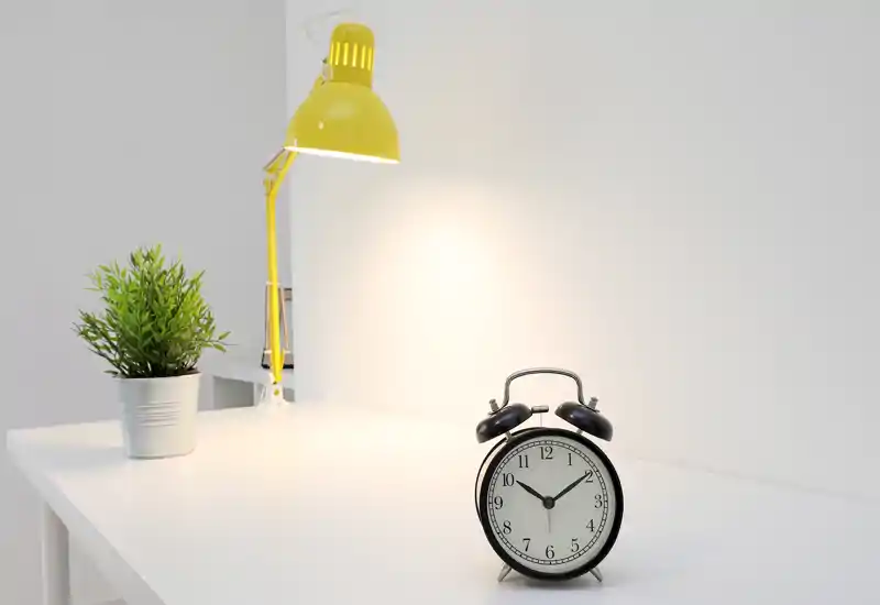 Scène de bureau minimaliste bien éclairée avec une lampe de bureau jaune, une petite plante en pot et un réveil classique sur une surface propre.
