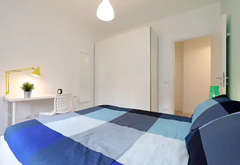 Bright, modern bedroom with a large double bed dressed in blue patchwork bedding, a white wardrobe, and a small desk with a lamp and plant — clean, minimal and well lit.