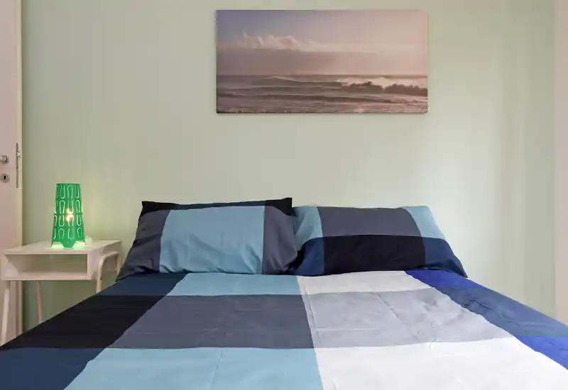 Bright, well-composed bedroom with a large double bed featuring blue check bedding, bedside table and a decorative lamp; calm wall art above the headboard enhances the relaxing atmosphere.