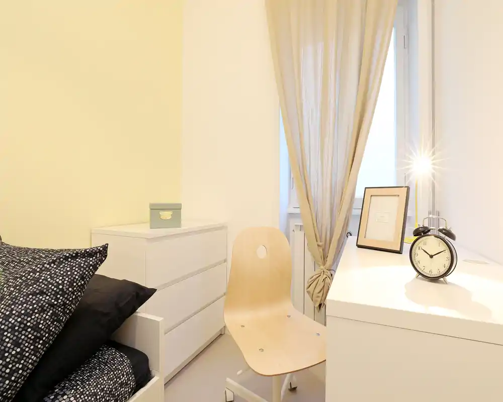 Coin chambre chaleureux et lumineux avec literie à motif, commode blanche et petit bureau près de la fenêtre muni d’un réveil rétro et d’un cadre.